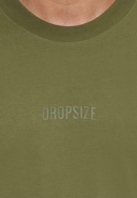 Olivinės spalvos medvilninė marškinė su klasikiniu apvaliu kaklu ir tekstūruotu "DROPSIZE" logotipu šiek tiek šviesesnėje atspalvyje. Lygi paviršius.