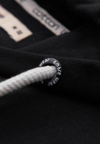 Abbigliamento in cotone nero con superficie texturizzata. Include un cappuccio con un cordino bianco intrecciato e un occhiello nero circolare con testo stampato.