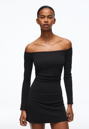 Une femme présente une mini-robe noire ajustée à manches longues et épaules dénudées sur un fond blanc uni.