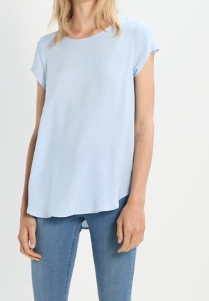 Kobieta ubrana w jasnoniebieską bluzkę z krótkim rękawem i niebieskie dżinsy, stojąca na białym, jednolitym tle.