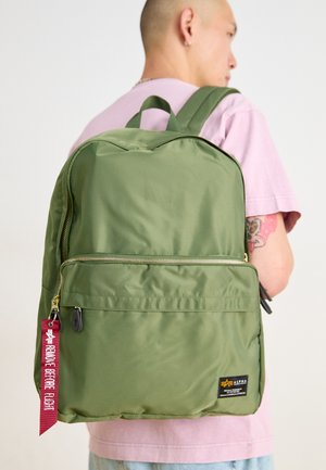 Green nylon backpack with a front zip pocket, gold hardware, and a red fabric tag reading "REMOVE BEFORE FLIGHT." Smooth texture, rounded shape.