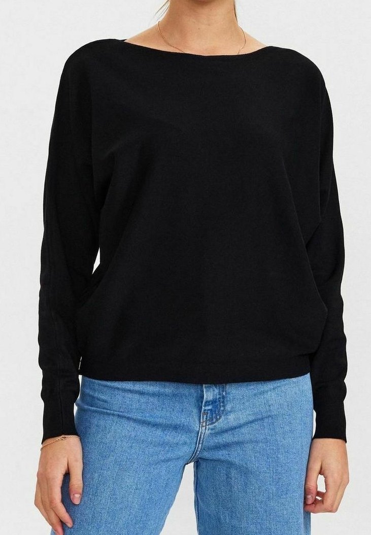 Femme portant un haut noir ample à manches longues et un jean bleu clair, debout avec les bras détendus le long du corps.