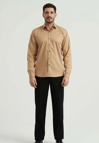 Chemise à manches longues marron clair avec un col boutonné et des boutons argentés, associée à un pantalon et des chaussures noires. Texture de tissu lisse.