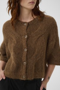 Γυναίκα που φοράει καφέ ζεστή πλεκτή ζακέτα με μεγάλα κουμπιά και μανίκια μέχρι το ν αγκώνα, συνδυασμένη με μαύρο παντελόνι.