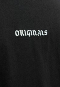 Melns T-krekls, kas izgatavots no mīksta kokvilnas, ar balti izšūtu tekstu "ORIGINALS" treknā, stilizētā fontā. Gluda tekstūra, pielāgota forma.