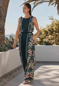 Bloemen jumpsuit in donkergroen met een ontspannen pasvorm. Heeft een gerimpelde taille, wijde benen en bloemenpatronen in roze en blauw aan de zijkanten.