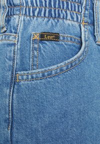 Modré džínové kalhoty mají smrštěný pas, zadní kapsu s viditelným švem a výraznou černou nášivku značky Lee.