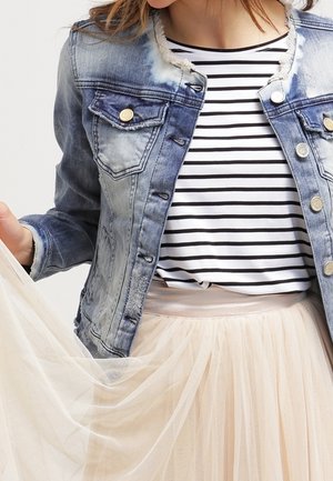 Frau trägt eine ausgewaschene blaue Jeansjacke über einem schwarz-weiß gestreiften Hemd und hält den durchscheinenden, beigen Tüllrock ihres Kleides.