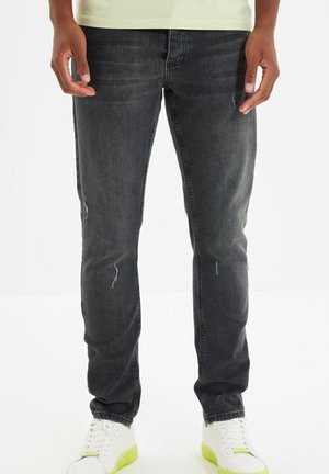 Uomo che indossa jeans neri skinny consumati e sneakers bianche con suola verde neon, in piedi con le mani rilassate lungo i fianchi.