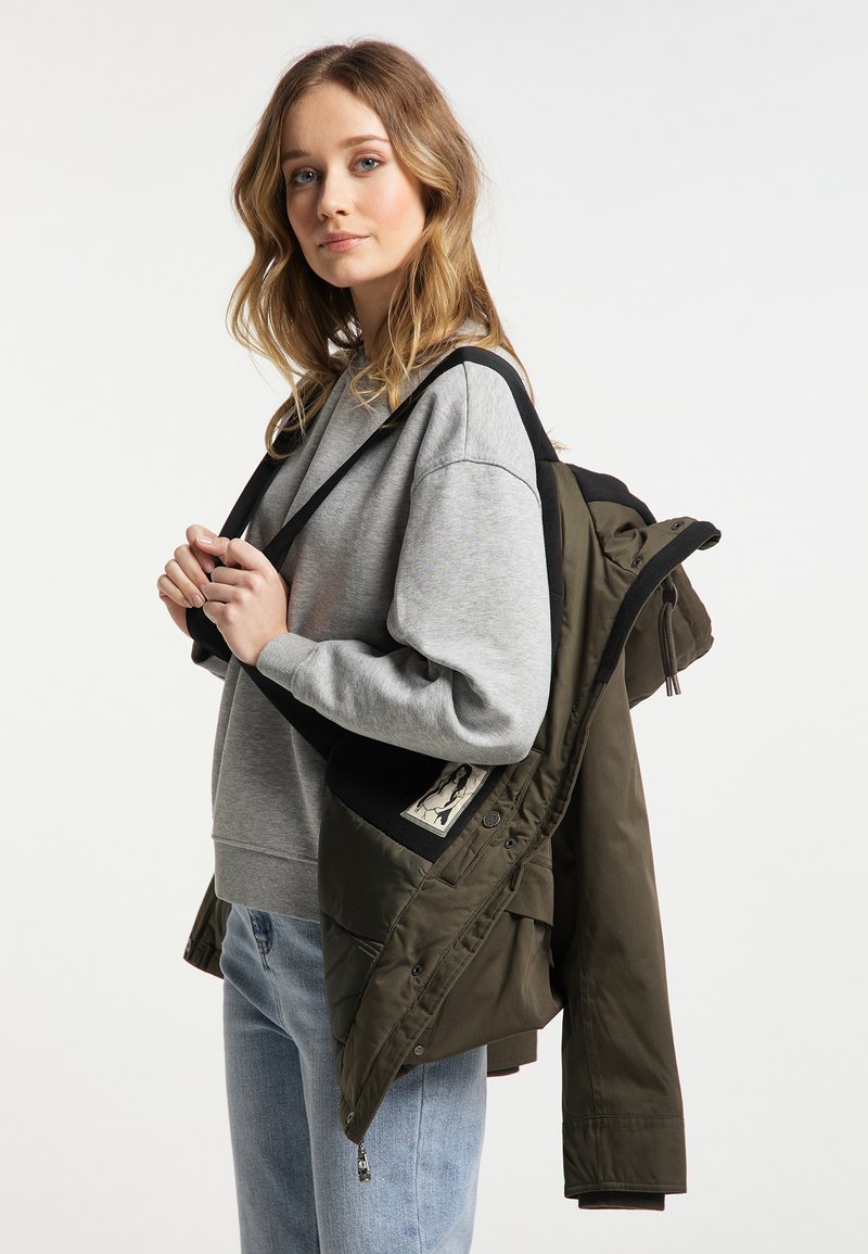 Olivgrüne Steppjacke mit schwarzem Akzent, über einem grauen Sweatshirt getragen. Verfügt über eine Kapuze und ein Etikett an der Jacke. Legere Jeans.