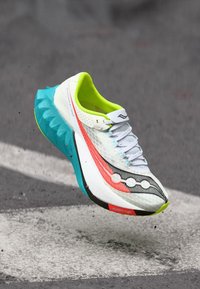 Weißer und neongrüner Laufschuh mit blauer Sohle in der Luft über Asphalt mit weißen Straßenmarkierungen.