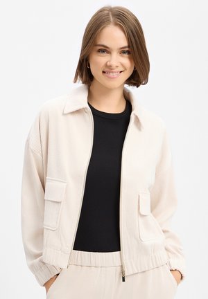 Junge Frau mit kurzen braunen Haaren, die eine beige Cordjacke über einem schwarzen Top trägt, lächelnd mit den Händen in den Jackentaschen.