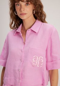 Camicia di lino rosa chiaro a maniche corte con colletto, caratterizzata da bottoni decorativi e un dettaglio ricamato sulla tasca. Texture liscia.