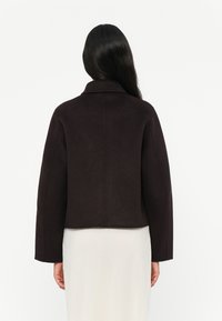 Brun cropped jakke i mykt stoff, med vide ermer og en enkel krage. Baksiden har et rent design uten synlige lukkinger.
