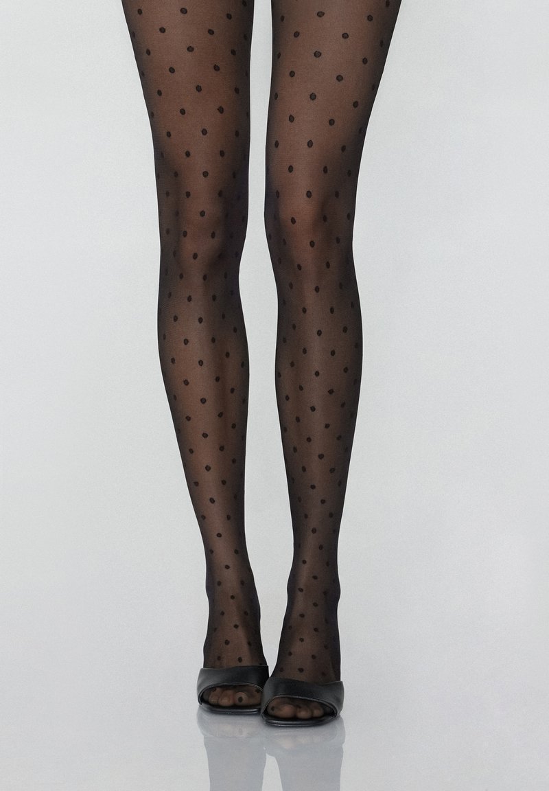 Collants noirs à pois avec une texture transparente ; petits pois ronds régulièrement espacés ; associés à des escarpins noirs ouverts à l'avant sur une surface lisse.