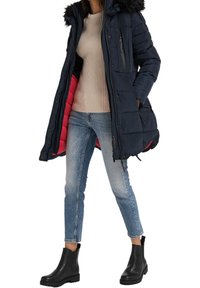 Marinblå vadderad jacka med huva av konstpäls, fickor med dragkedja, röd insida; matchad med en beige ribbad tröja, ljusblå jeans och svarta ankle boots.