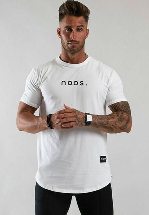 Muskelös man med tatueringar på höger arm, iklädd en vit t-shirt med texten "noos.", svarta byxor, smartklocka och svart pärlarmband, står mot en enfärgad bakgrund.