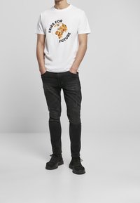 Muž oblečený v bílé tričko s glóbem ve tvaru hranolků a nápisem "Hranolky pro budoucnost", černé džíny a černé tenisky, stojící s rukama v kapsách.