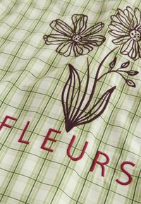 Tissu à carreaux verts et blancs avec des fleurs brunes brodées et des lettres rouges formant le mot "FLEURS".
