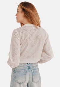Weiße Bluse mit aufwändigem Durchbruchsdesign, langen Ärmeln und rundem Ausschnitt, kombiniert mit hellblauen Jeans.