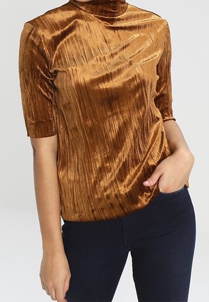 Haut à col montant à manches courtes en tissu brillant orange brûlé avec des plis verticaux, assorti à un pantalon en jean denim sombre.