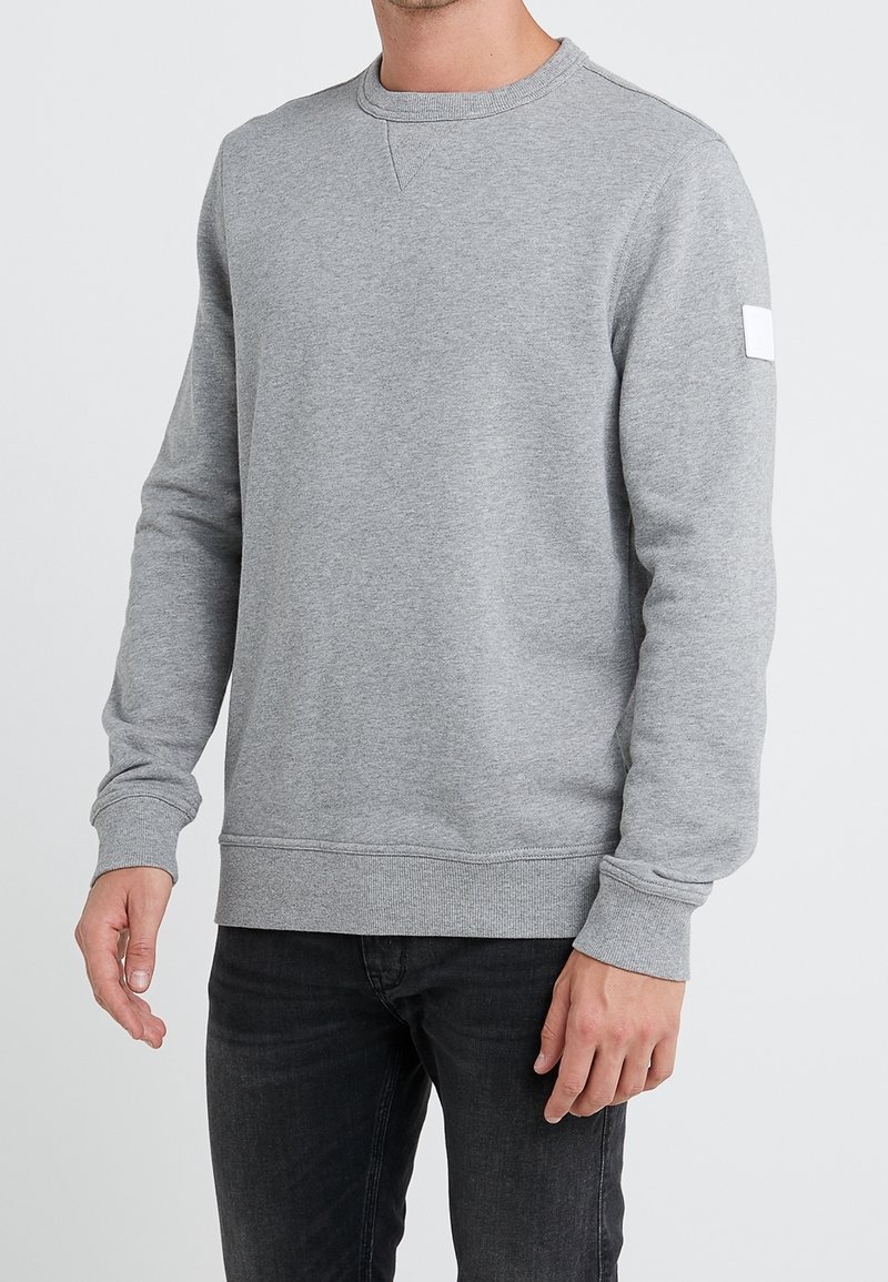 Homme portant un sweat-shirt à manches longues gris clair uni avec poignets et ourlet côtelés, associé à un jean noir, debout devant un fond uni.
