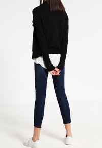 Camiseta negra de manga larga con un respaldo drapeado sobre una camisa blanca, combinada con jeans ajustados oscuros y zapatillas blancas, vista desde detrás.