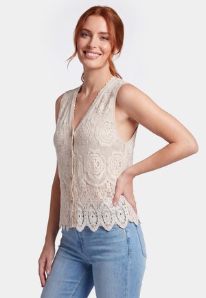 Lächelnde Frau mit roten Haaren trägt ärmelloses beiges florales Spitzenoberteil und hellblaue Jeans, posiert mit einer Hand an der Hüfte.