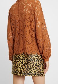 Frau trägt ein burnt orange Spitzenoberteil mit langen Ärmeln und Knöpfen sowie einen kurzen Rock im Leopardenmuster, von hinten gezeigt.