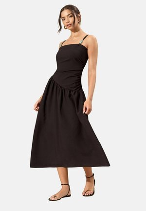 Frau trägt ein ärmelloses schwarzes Midikleid mit mit Perlen verzierten Trägern und flachen schwarzen Sandalen, steht vor einem schlichten weißen Hintergrund.