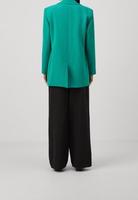 Blazer turquesa de manga larga, con una abertura en la espalda, combinado con pantalones de pierna ancha negros. Tejido suave sin patrones o detalles visibles.