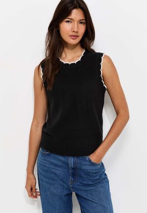 Giovane donna con lunghi capelli scuri che indossa un top nero senza maniche in maglia con bordatura bianca e jeans blu, in piedi con una mano in tasca.