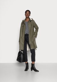 Impermeabile verde oliva con cappuccio regolabile, bottoni frontali e tasche laterali, indossato sopra un maglione grigio e jeans neri. Stivali neri alla caviglia e borsa.