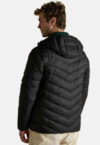 Muž s kučeravými vlasmi, oblečený v čiernej prešívanej kapucňovej bunde a svetlých nohaviciach, stojí bokom proti jednofarebnému pozadiu.