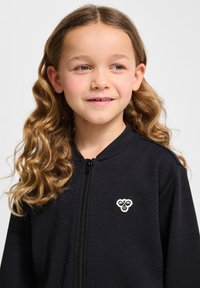 Svart zip-up jacka i mjukt tyg, med rund halsringning och en broderad logotyp på bröstet.