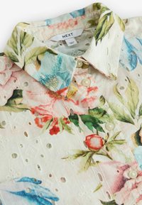 Blommig skjorta med vit bas, med färgglada blommönster. Volangdetaljer vid ögonen, knäppning med knappar och spetskrage.