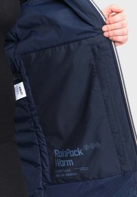 Unutrašnjost mornarsko plave puffer jakne s oznakom "RainPack Warm." Sadrži glatku teksturu, simbole za vodootpornost i sklopivi dizajn za dodatnu toplinu.