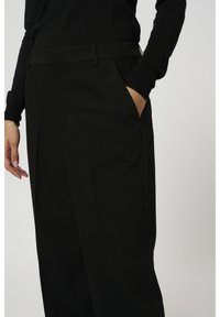 Personne portant un pantalon noir et un haut à manches longues noir, la main droite dans la poche et le bras gauche détendu le long du corps, sur un fond uni.