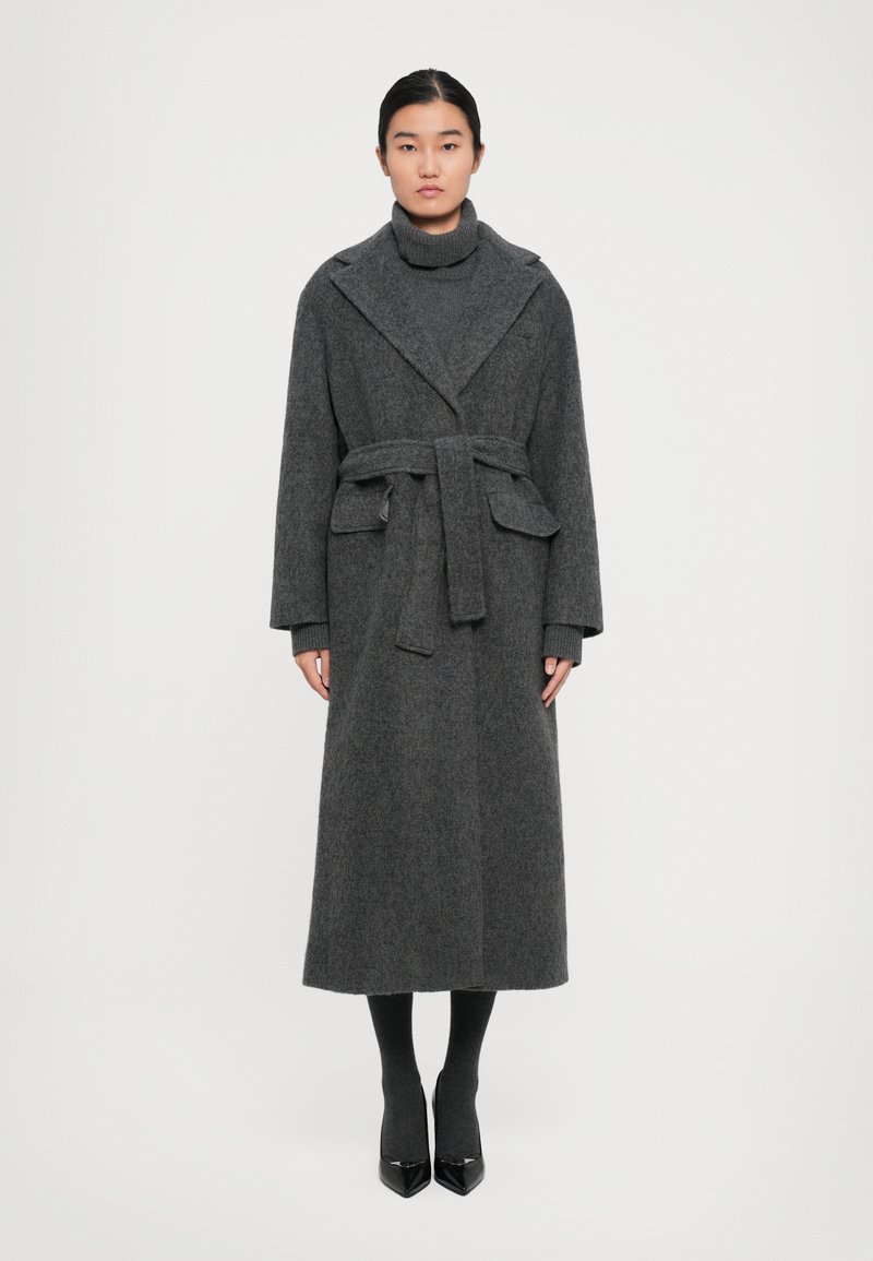 Long manteau en laine grise avec une ceinture, grand col et poches latérales ; associé à un pull col roulé gris et des escarpins noirs à bout pointu.