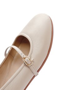 Beige leather ballet flat with a round toe, ankle strap, and gold-tone buckle detail; smooth texture and minimal stitching accents.
