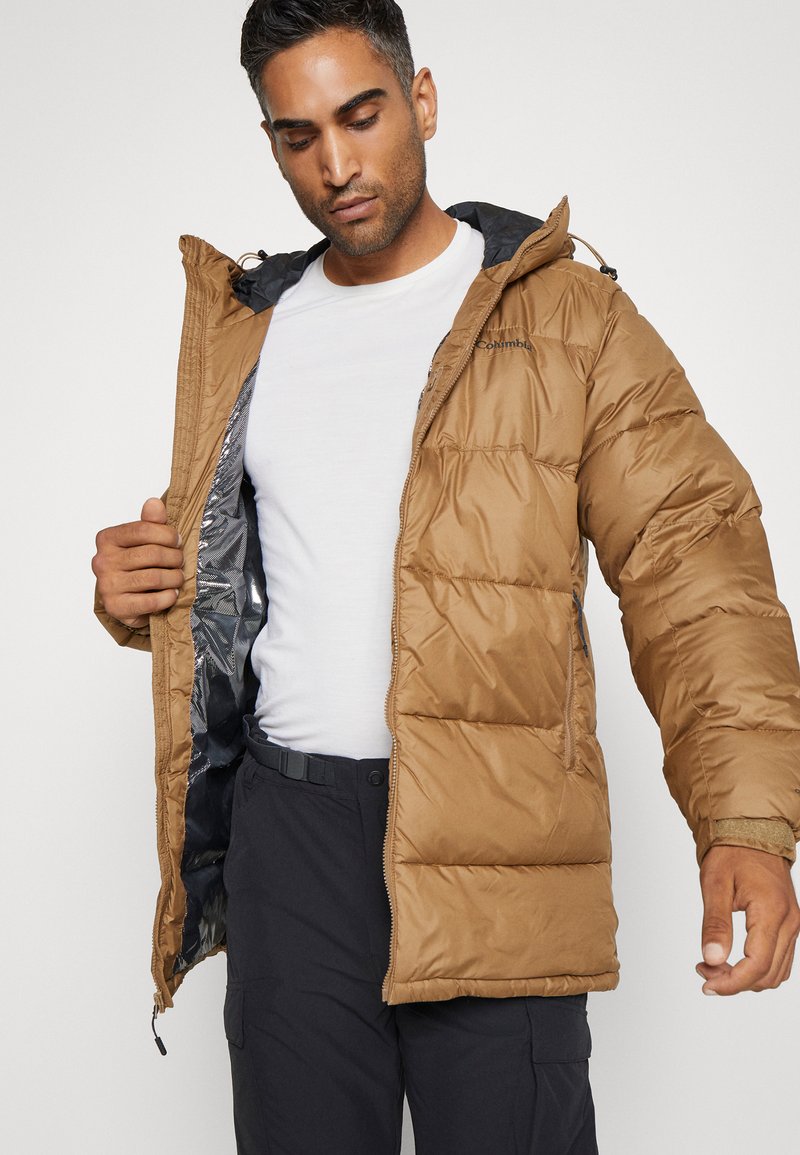 Jasnobrązowa pikowana kurtka z zamkiem z przodu, kapturem i błyszcząco-srebrną podszewką, noszona na białej koszuli i ciemnych spodniach.