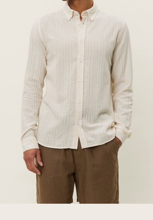 Homme portant une chemise blanche texturée boutonnée avec col et manches longues, associée à un pantalon marron ample, debout avec une main dans la poche.
