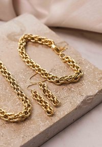 Gouden metalen sieraden set met een chunky schakelarmband, bijpassende oorbellen en een ketting met onderling verbonden lussen, tentoongesteld op een stenen oppervlak.