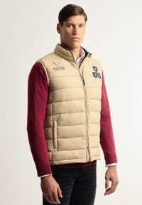 Chaleco acolchado beige con cremallera frontal, bolsillos laterales y logo. Usado sobre una camiseta de manga larga roja. Presenta tres insignias bordadas en el pecho.