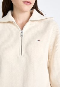 Kobieta ubrana w kremowy sweter z ribbowanym splotem, z kołnierzem na zamek błyskawiczny oraz małym haftowanym logo w kolorach czerwonym, białym i czarnym na piersi.