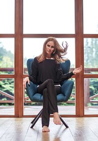 A blue upholstered armchair with a sleek design, complemented by a wooden floor. A person in a black top and pants sits relaxed, barefoot.