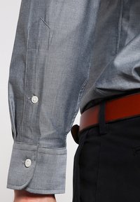 Zblízka záběr na šedý rukáv trička s dlouhým rukávem a bílými knoflíky, nošený s černými kalhotami a hnědým koženým páskem.