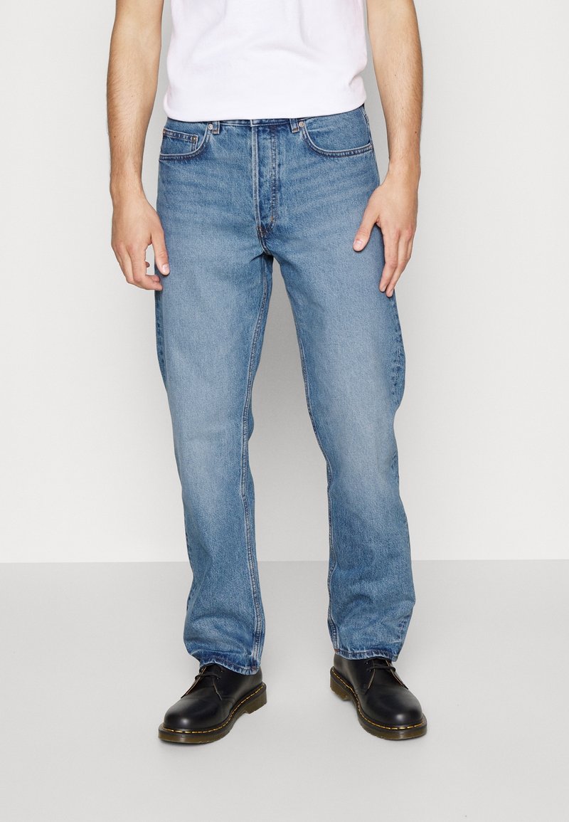 Homme portant un jean droit bleu, des chaussures noires à lacets et un t-shirt blanc, debout devant un fond uni.