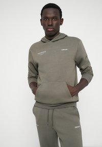 Sweatshirt de cor verde-oliva e calças combinando, feitas de tecido macio, com um bolso à frente, punhos elásticos e detalhes impressos em branco.