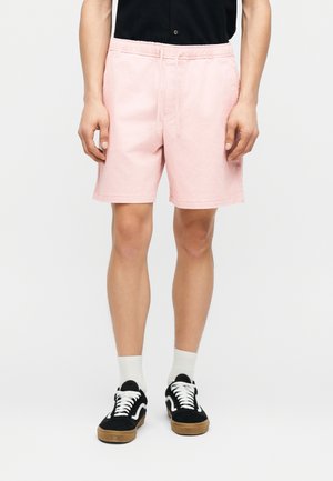 Person, die hellrosa Kordelzugshorts, schwarze Sneaker mit weißen Schnürsenkeln, weiße Socken und ein schwarzes Hemd mit Knöpfen trägt und steht.